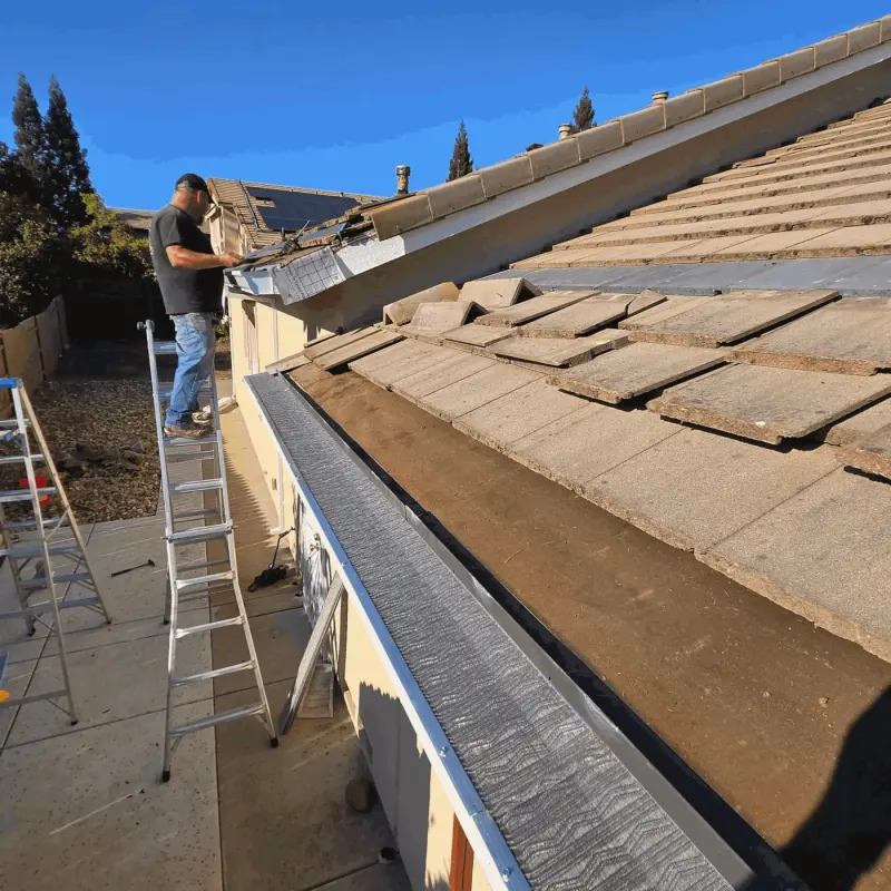 Installer on a ladder fitting gutter guard while roof tiles are lifted to tuck the guard underneath