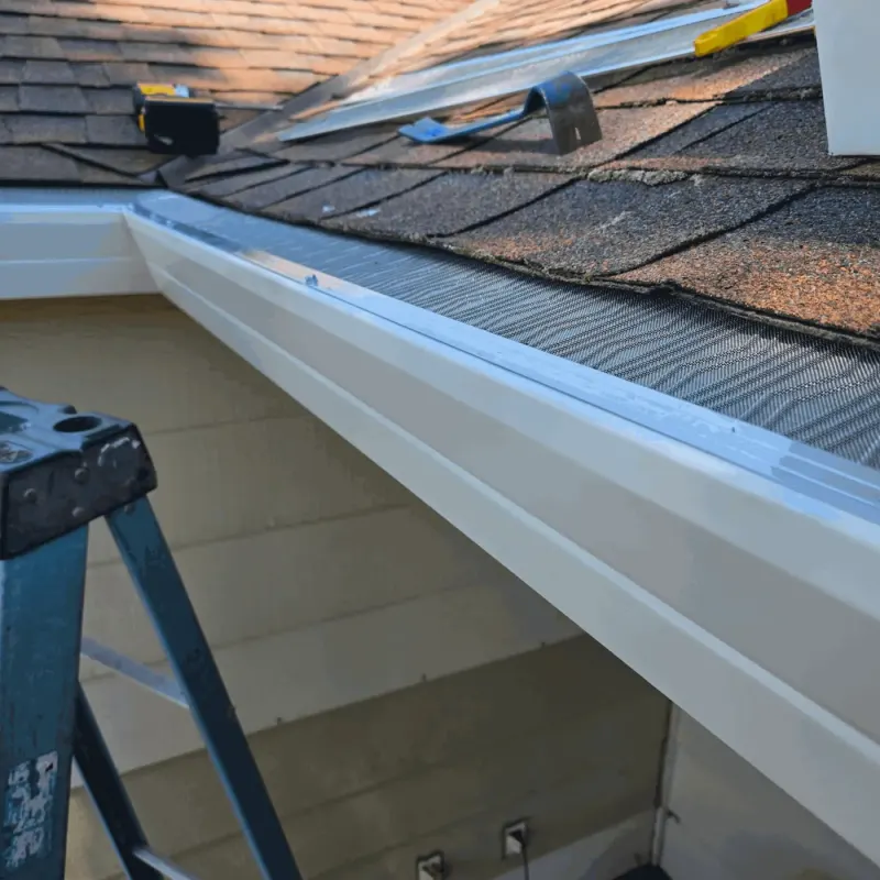 Micro-mesh gutter guard going onto a white seamless gutter on a composition shingle roof, guard panels and tools staged on the roof