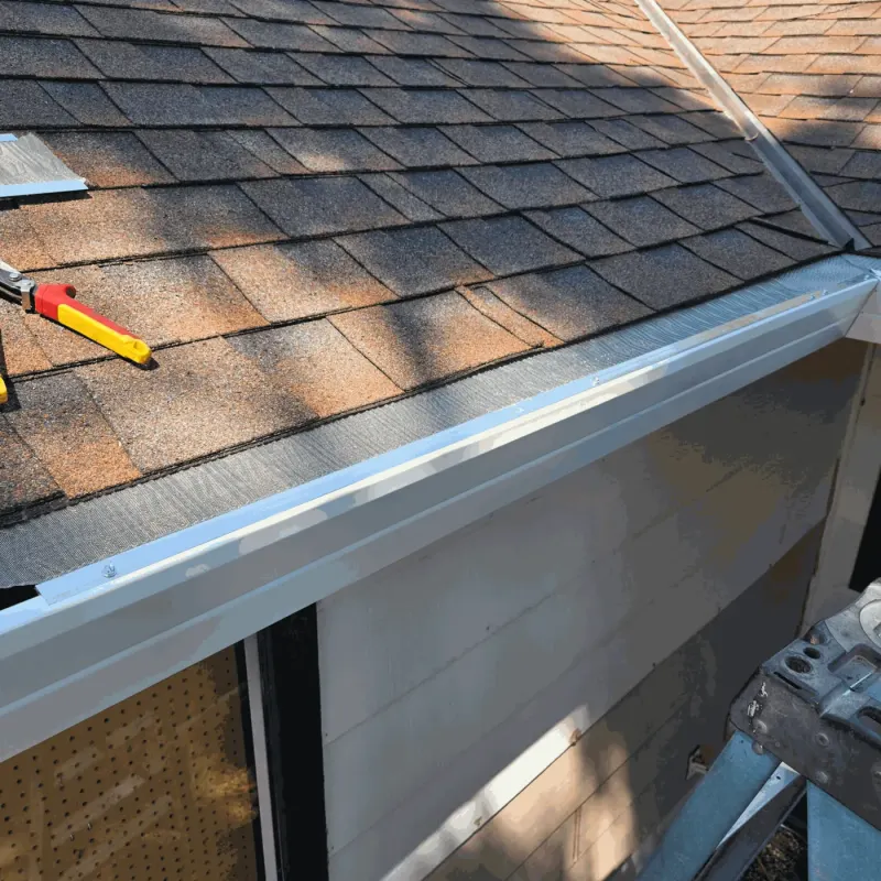 Stainless micro-mesh gutter guard screwed to the front lip of a white seamless gutter on a shingle roof